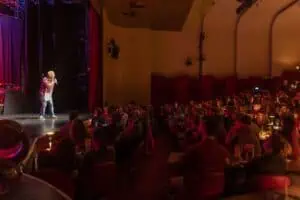 Ein Komiker tritt auf einer schwach beleuchteten Bühne auf und fesselt das Publikum im Theater, ähnlich wie bei einem mitreißenden Poetry Slam.